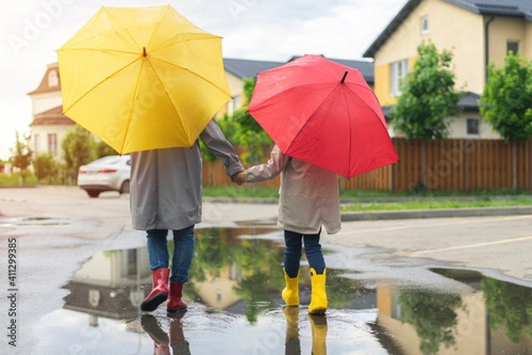 Fototapeta Mom and daughter rainy weather go home under umbrellas and in rubber boots. mother and child walk down the street in autumn through puddles