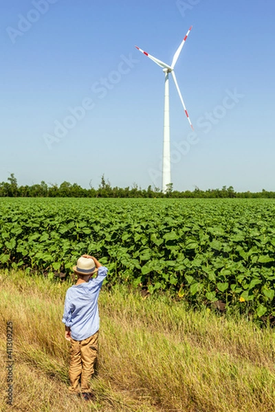 Fototapeta A child from a dirt road looks at wind turbines against a background of cloudy blue sky and green field