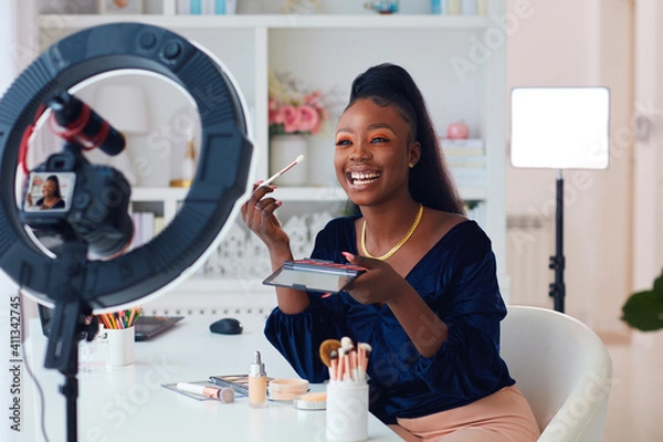 Obraz happy young african american woman streaming a beauty vlog from home, online content creator applying a makeup on