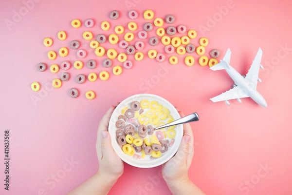 Obraz Colorful fruit corn rings with milk in the bowl, children's hands, airplane on a pink background. Children's breakfast option with cornflakes.