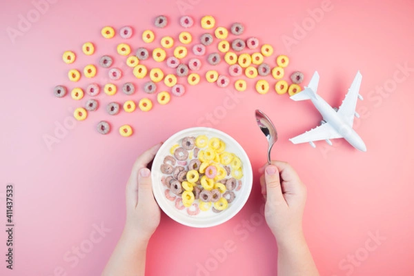 Obraz Colorful fruit corn rings with milk in the bowl, children's hands, airplane on a pink background. Children's breakfast option with cornflakes.