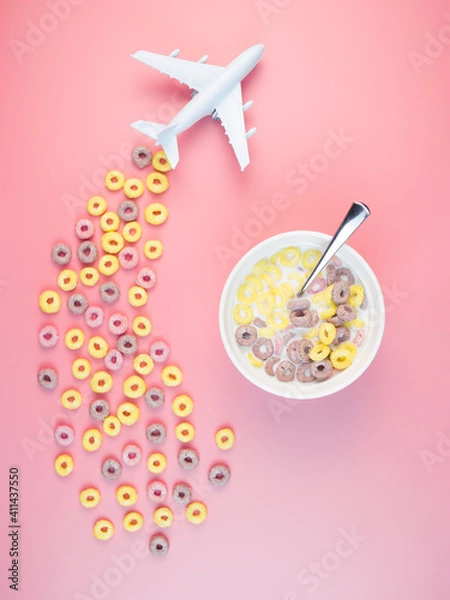 Obraz Colorful fruit corn rings with milk in the bowl, airplane on a pink background. Children's breakfast option with cornflakes.