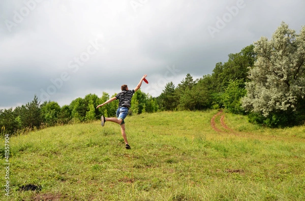Fototapeta A teenager in a green meadow catches a flying disc in a jump