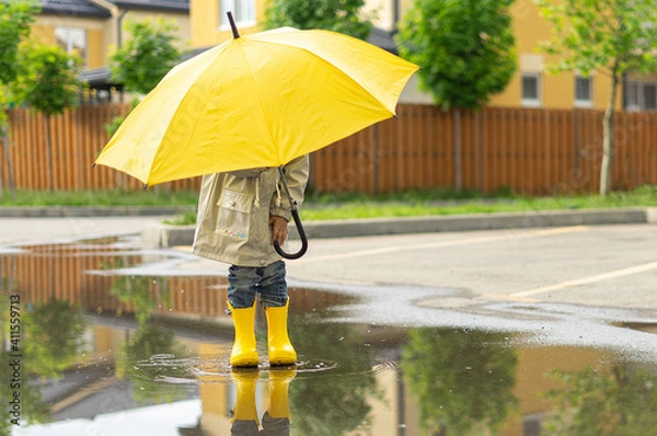 Fototapeta A child holds a yellow umbrella standing in a puddle outside. On sunny day happy kid walks near the house, plays, has fun