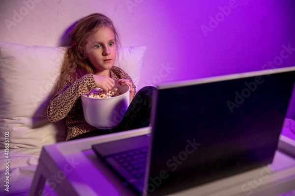 Obraz Little girl watching cartoon on laptop in bed in the evening and eating popcorn. She is surprised looking at the screen. Home entertainment. Evening twilight