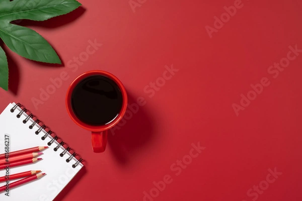 Fototapeta Desk top view with red coffee cup, pencil and notebook on red background.