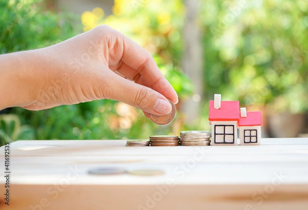 Fototapeta Hand holding coin with miniature house in concept of saving money for buying home, real estate or financial