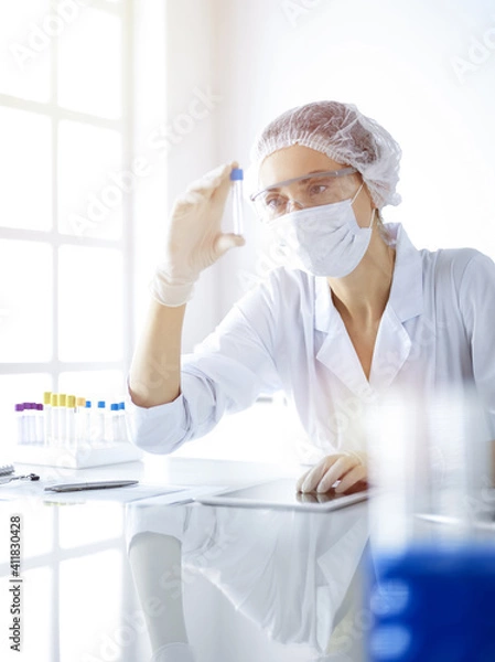Fototapeta Professional female scientist in protective eyeglasses researching tube with reagents in sunny laboratory. Medicine and science researching