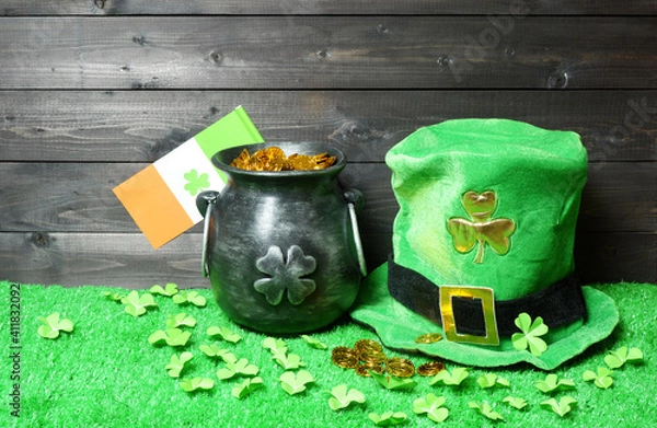 Fototapeta Cast iron pot with treasure, irish flag with clover leaf and leprehaun hat, clover leaves and coins on green grass, dark wooden planks background. Saint Patricks Day banner, poster, flyer, invitation