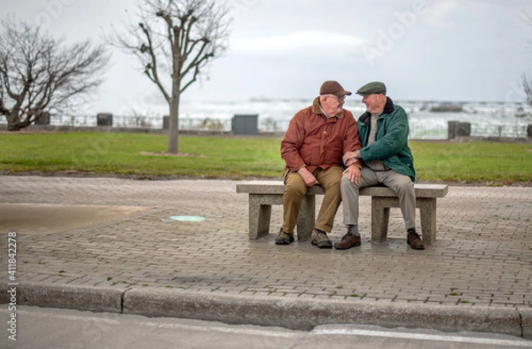 Fototapeta Senior Caucasian gay married couple sitting on a bus bench, holding hands whilst chatting and smiling.