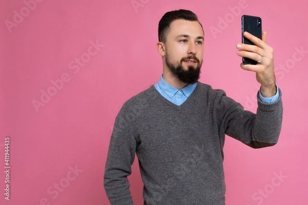 Fototapeta Handsome young man wearing casual stylish clothes standing isolated over background wall holding smartphone taking selfie photo looking at mobile phone screen display