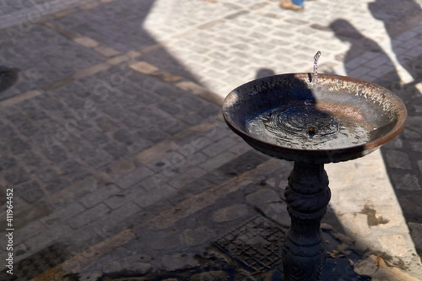 Fototapeta Fuente funcionando entre el sol y la sombra con un empedrado de la calle de fondo