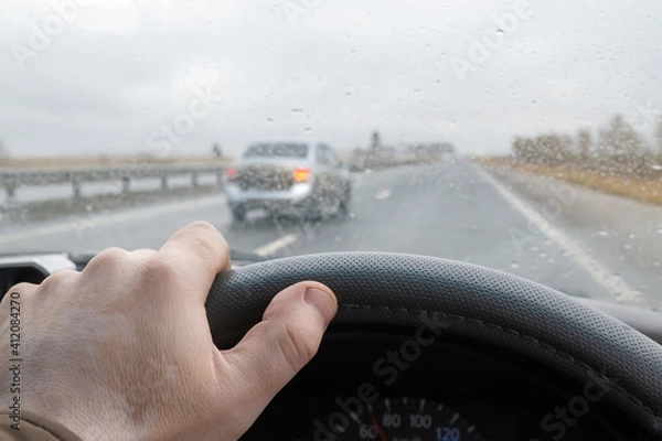Fototapeta view of the driver's hand on the steering wheel of a car that is driving on the highway and in front of it an overtaking car makes a maneuver of changing lanes in rainy cloudy weather