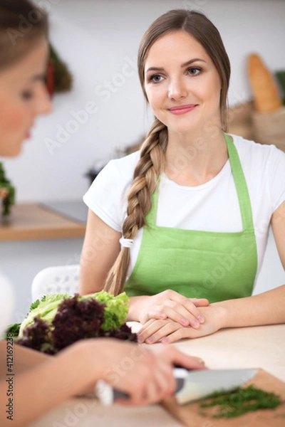 Fototapeta Two women are cooking fresh salad in a kitchen and having a pleasure talk. Friends and Chef Cook concept