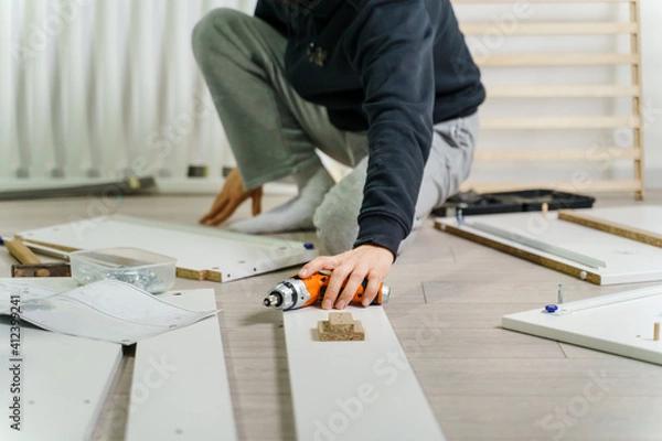 Fototapeta One unknown Man alone Putting Together Self Assembly Furniture sitting on the floor at Home holding electric screwdriver looking the instructions - side view DIY concept real people copy space