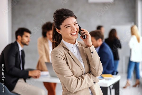 Fototapeta Smiling cheerful young businesswoman standing in boardroom and talking on the phone. In background are her colleagues working on important project.