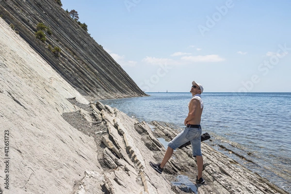 Obraz Summer sea landscape. The guy walking along the stone wild beach stopped and looks up. On the horizon, clear blue sky and steep cliffs. The resort city of Gelendzhik. Russia, Black Sea coast