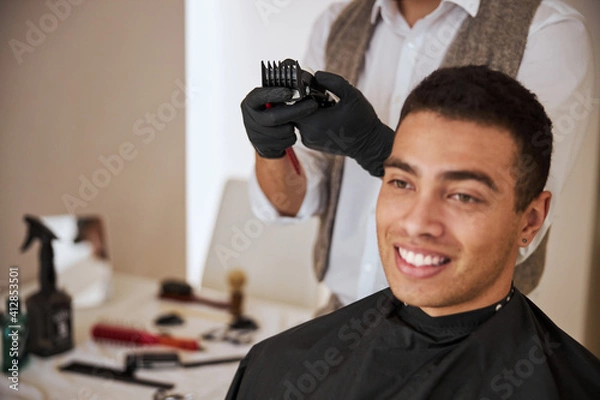 Obraz Unrecognized barber in gloves holding electrical trimmer behind the client in room indoors