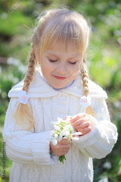 Fototapeta Cute little girl considering a fresh bouquet of snowdrops. Spring time. Little girl in white walks in the forest