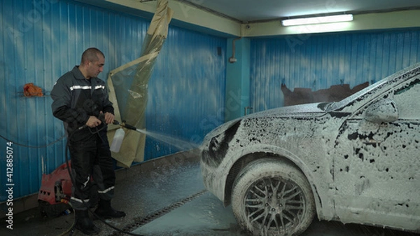Fototapeta A Caucasian man works in a car wash service. A man in a worker's uniform sprays a car with detergent