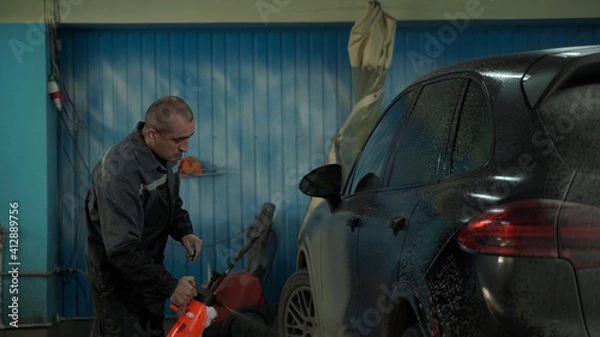 Fototapeta A Caucasian man works in a car wash service. A man in a worker's uniform washes the car thoroughly by spraying it with a water pistol.