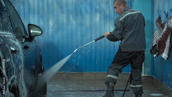 Fototapeta A Caucasian man works in a car wash service. A man in a worker's uniform washes the car thoroughly by spraying it with a water pistol.