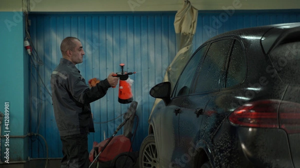 Fototapeta A Caucasian man works in a car wash service. A man in a worker's uniform washes the car thoroughly by spraying it with a water pistol.