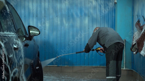 Fototapeta A Caucasian man works in a car wash service. A man in a worker's uniform washes the car thoroughly by spraying it with a water pistol.