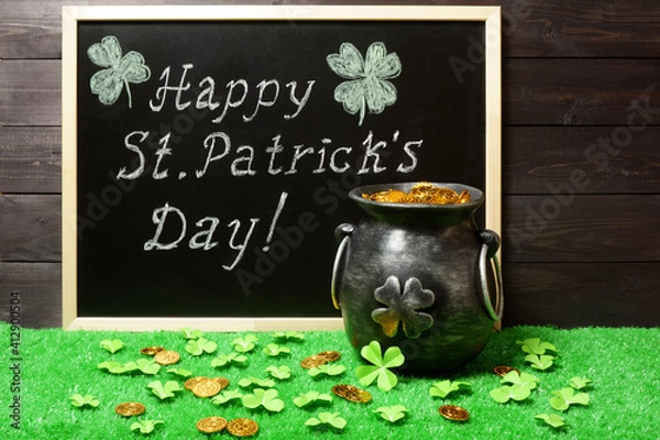 Fototapeta Banner, poster, invitation template. Happy St. Patricks Day chalk lettering on blackboard, cast iron pot full of leprechaun golden treasure, clover leaves and coins on green grass, dark wood bg