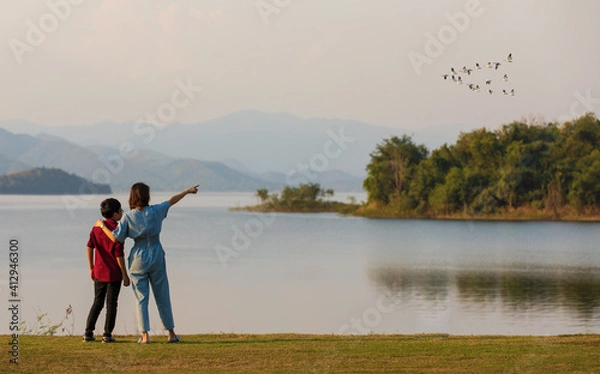 Fototapeta mother and son standing beside big lake and see mountain view in the background, mom pointing finger to birds flying in sky. Idea for family tourist travels together