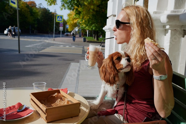 Fototapeta A 40-year-old blonde woman on the street at a table drinks coffee, next to her is a Cavalier King Charles Spaniel dog in a comic situation