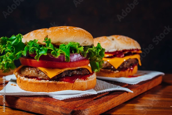 Fototapeta Delicious burgers with Bacon and cheddar cheese and with lettuce, tomato and red onion and bacon and cheddar on homemade bread with seeds and ketchup on a wooden surface and black background.