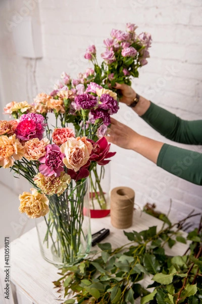 Obraz Florist's workplace. Tools and flowers before starting work on the table against the background of a white brick wall. A florist chooses a flower for a bouquet. Tutorial. Step by step