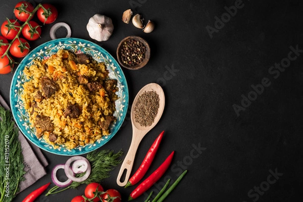 Fototapeta A large plate filled with pilaf, next to cherry tomatoes, fresh pepper . garlic, zeros and a mixture of peppers. All on a dark background.