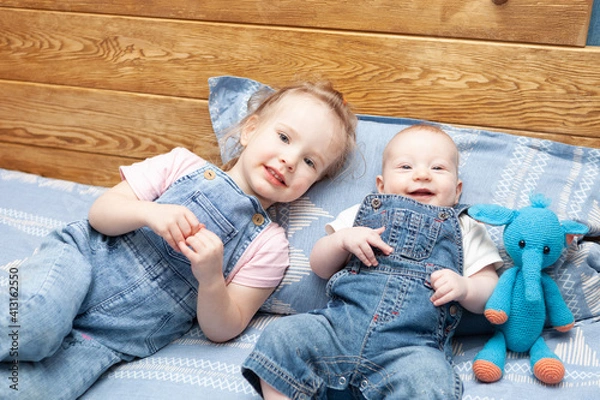 Fototapeta two smiling little children (brother and sister) in denim suits lie on the bed 