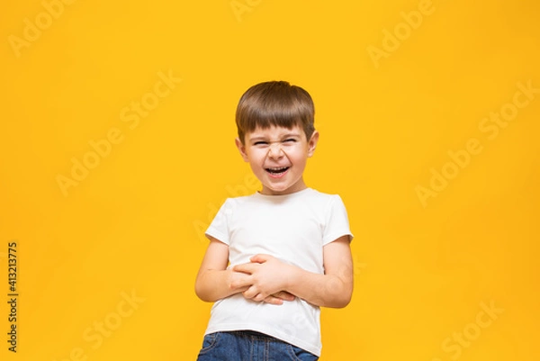 Fototapeta Cheerful boy laughs with his mouth wide open in a white T-shirt holding his hands on his stomach. on an isolated yellow background