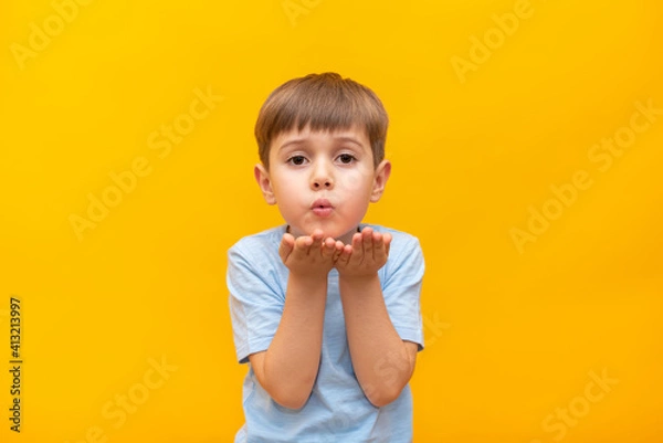 Fototapeta Attractive little boy blowing a kiss to the camera, demonstrating love for his girlfriend or saying goodbye from a distance, isolated on yellow studio background. 