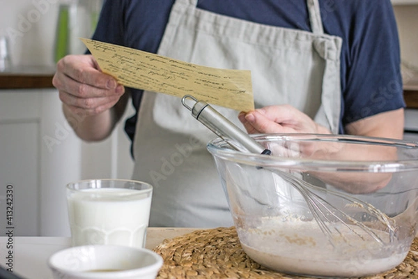 Fototapeta A man without a face in an apron kneads dough for homemade bread according to a recipe written on a piece of paper, which he holds in his hands. Cooking at home learning concept, authentic hobby