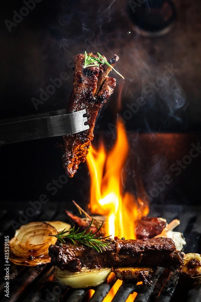 Fototapeta Grilled pork ribs on the grill chopped on a fork against a fire. The cook holds a fork in his hand. concept cooking meat. Close up. place for text