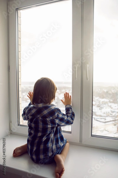 Fototapeta child boy sits on a window sill in winter on a high floor