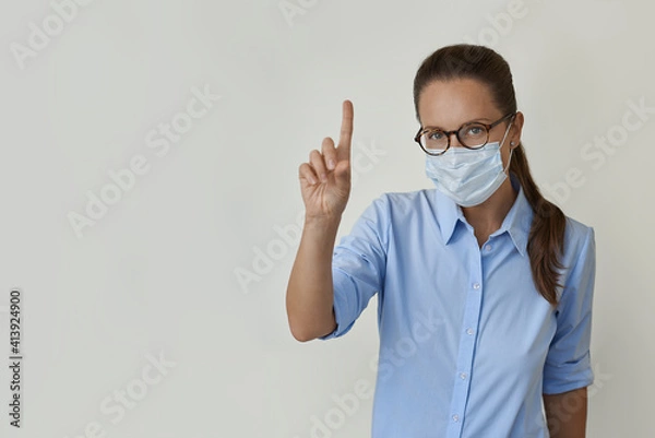 Fototapeta On the right side of the image is a woman wearing a medical face mask and a blue shirt, pointing her finger at an empty space