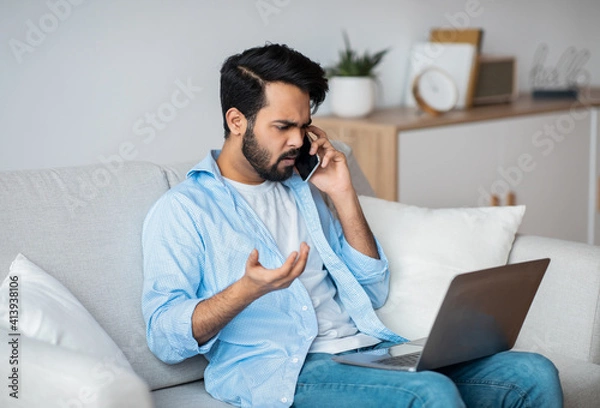 Fototapeta Computer Problems. Frustrated arab guy using cellphone and looking on laptop screen