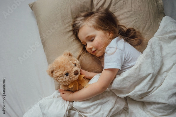Fototapeta Little girl sleeping in bed embracing soft toy at home, top view