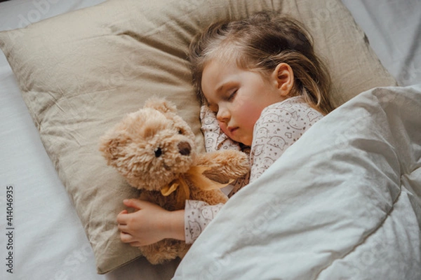 Fototapeta Little girl sleeping in bed embracing soft toy at home, top view