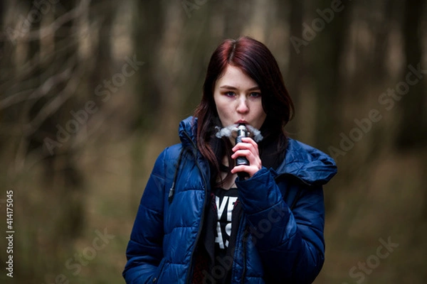 Fototapeta Vape teenager. Young pretty caucasian brunette girl with problem skin smoking an electronic cigarette on the street in the spring. Deadly bad habit.
