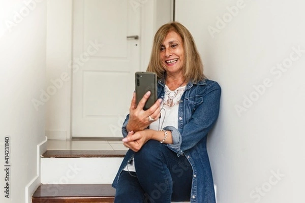 Obraz Happy mature old 60s woman holding smartphone using mobile phone app for video call, laughing while watching funny video, feeling excited winning online lottery bid on cellphone sits on couch at home.