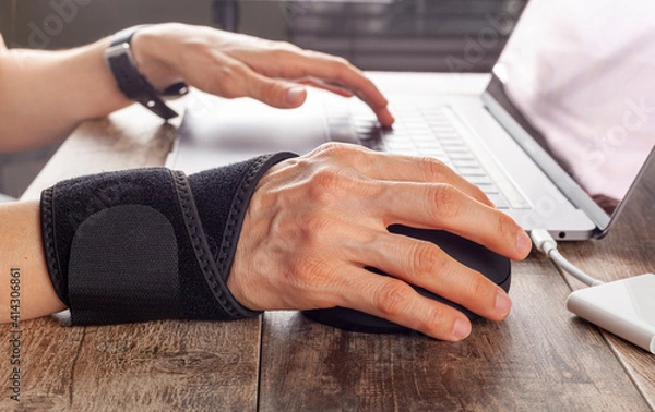 Fototapeta Chronic trauma to the wrist joint  in people using computer mouse may lead to disorders that cause inflammation and pain. A woman working on desk uses wrist support brace and ergonomic vertical mouse