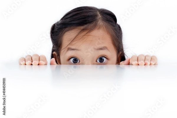 Fototapeta Portrait of an Asian girl peeking through the surface of table isolated on white background.