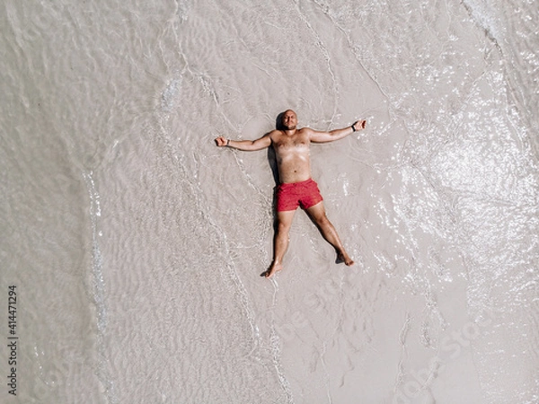 Fototapeta Top view of a satisfied man in red shorts lying on the hot sand of the beach and sunbathing under the rays of the hot sun. Summer concept.