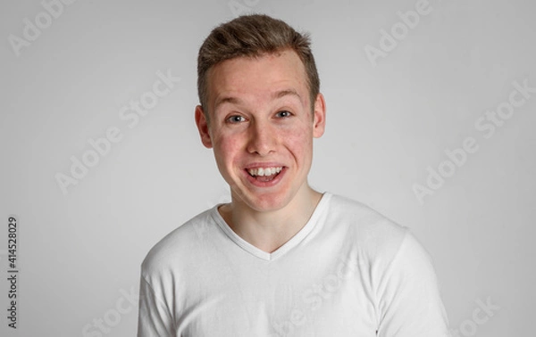 Fototapeta teen guy looking into the frame happy surprised can't believe raised eyebrows
 and smiling with teeth on a white background in a white T-shirt